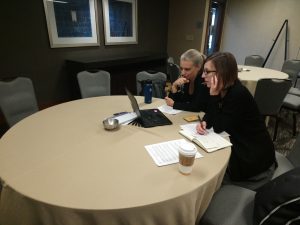 duas mulheres sentadas com mesa à frente, olhando o computador e anotando